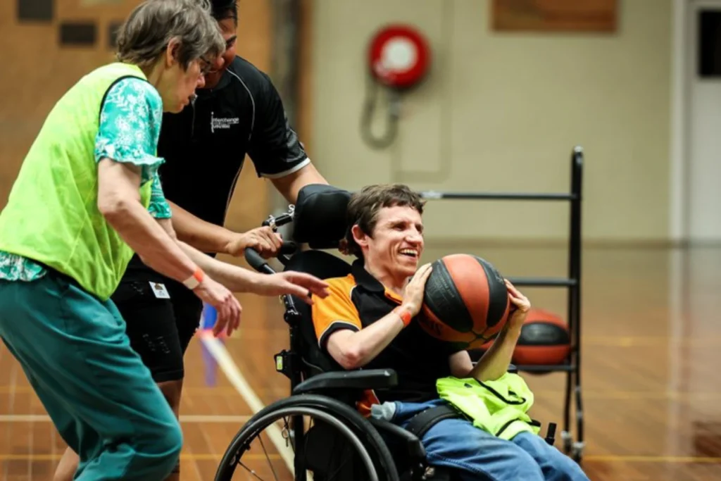 A man in a wheelchair smiling and holding a basketball, supported by two people during an indoor sports session