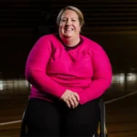 A smiling woman in a bright pink top sitting in a wheelchair on a sports court.