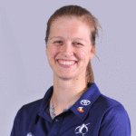 A smiling woman wearing a navy sports polo shirt with logos, standing against a light grey background.