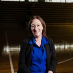 A woman wearing a blue shirt and black jacket, smiling while seated on an indoor sports court.