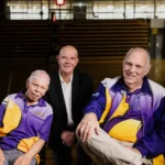 Three men smiling on an indoor sports court, two wearing purple and yellow sports jackets, one in a black suit