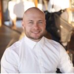 A man with a shaved head and beard, smiling, wearing a white button-up shirt, seated indoors with blurred background elements.