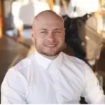 A man with a shaved head and beard, smiling, wearing a white button-up shirt, seated indoors with blurred background elements.