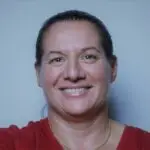A woman with dark hair tied back, smiling, wearing a red top and a gold necklace, against a plain light blue background.