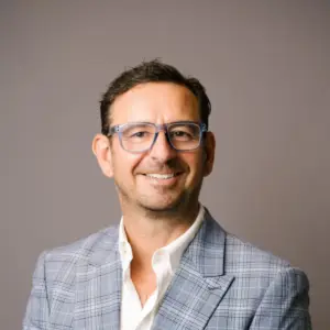 Portrait of a smiling man wearing blue-framed glasses, a light grey check blazer, and a white shirt, facing the camera against a neutral background.