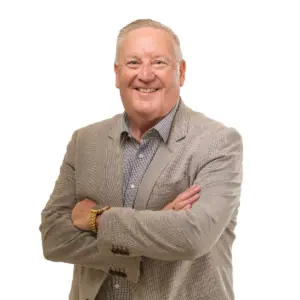 Portrait of a smiling man with short grey hair, wearing a beige blazer over a checkered shirt, standing with his arms crossed against a plain light background.