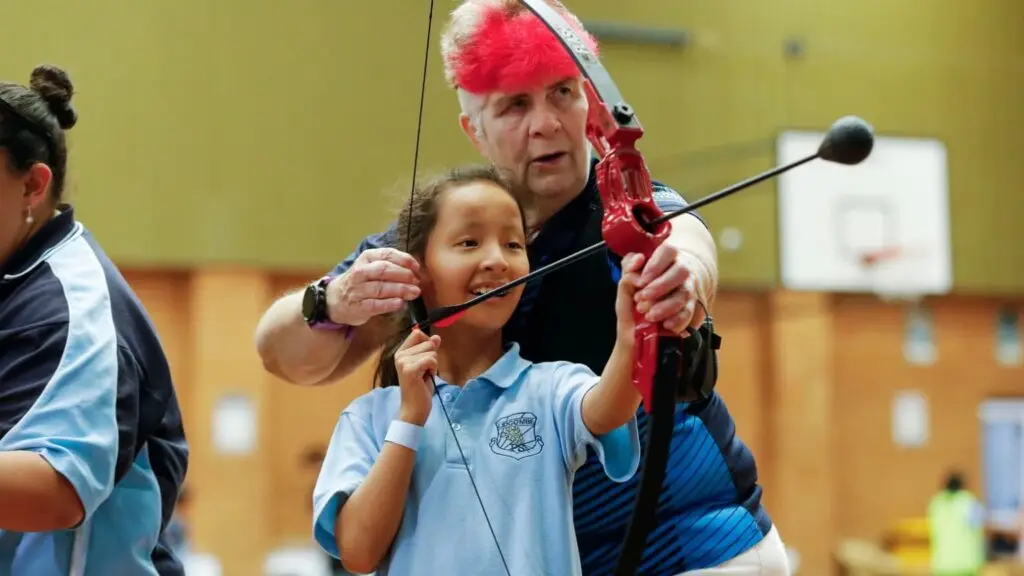 Child with disability smiling while holding archery bow alongside a coach during an indoor sport activity, with other participants in the background.