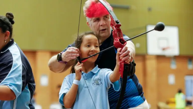 Child with disability smiling while holding archery bow alongside a coach during an indoor sport activity, with other participants in the background.