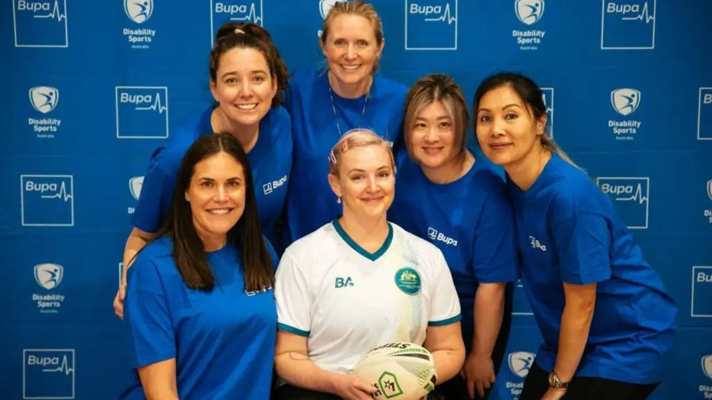 Group of people, including a person with disability seated at the front, smiling for a photo in front of a blue Disability Sports Australia backdrop with Bupa logos.