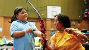 Child with disability participating in an inclusive sport activity archery indoors, holding equipment and engaging with a coach while other participants take part in the background.