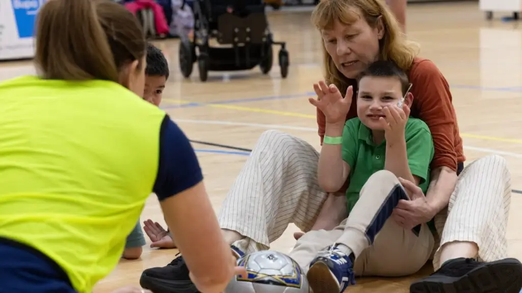 Child with disability sitting on the floor during an inclusive sport activity, smiling and raising their hands while supported by an adult, with a coach engaging them using a ball in an indoor sports setting.
