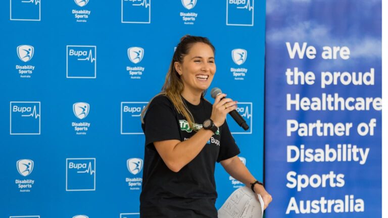 Presenter standing in front of a blue backdrop with Disability Sports Australia and Australian Sports Commission logos, with text reading: “We are the proud National Health Partner of Disability Sports Australia.”