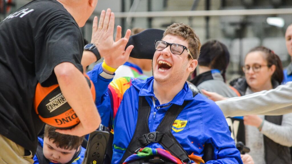 Child with disability smiling and raising their hands during a group sport activity, with other participants and volunteers in the background.