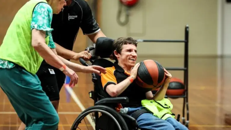 A man in a wheelchair smiling and holding a basketball, supported by two people during an indoor sports session
