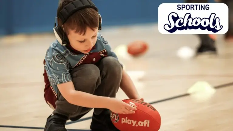 Young child wearing noise-cancelling headphones crouched on an indoor court, focusing on placing a small marker on a red AFL football, with cones and activity equipment in the background and a Sporting Schools logo in the top corner.